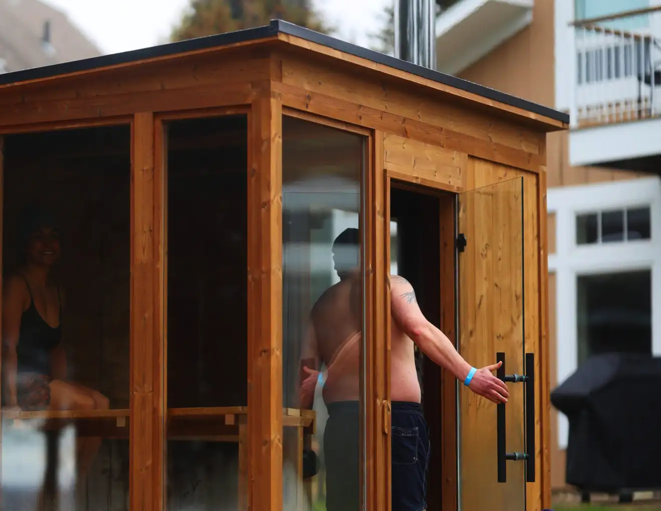 Side view of a man with swim trunks and a blue wrist band standing halfway out the door of a Traditional Mod Pod Sauna with full-height windows.