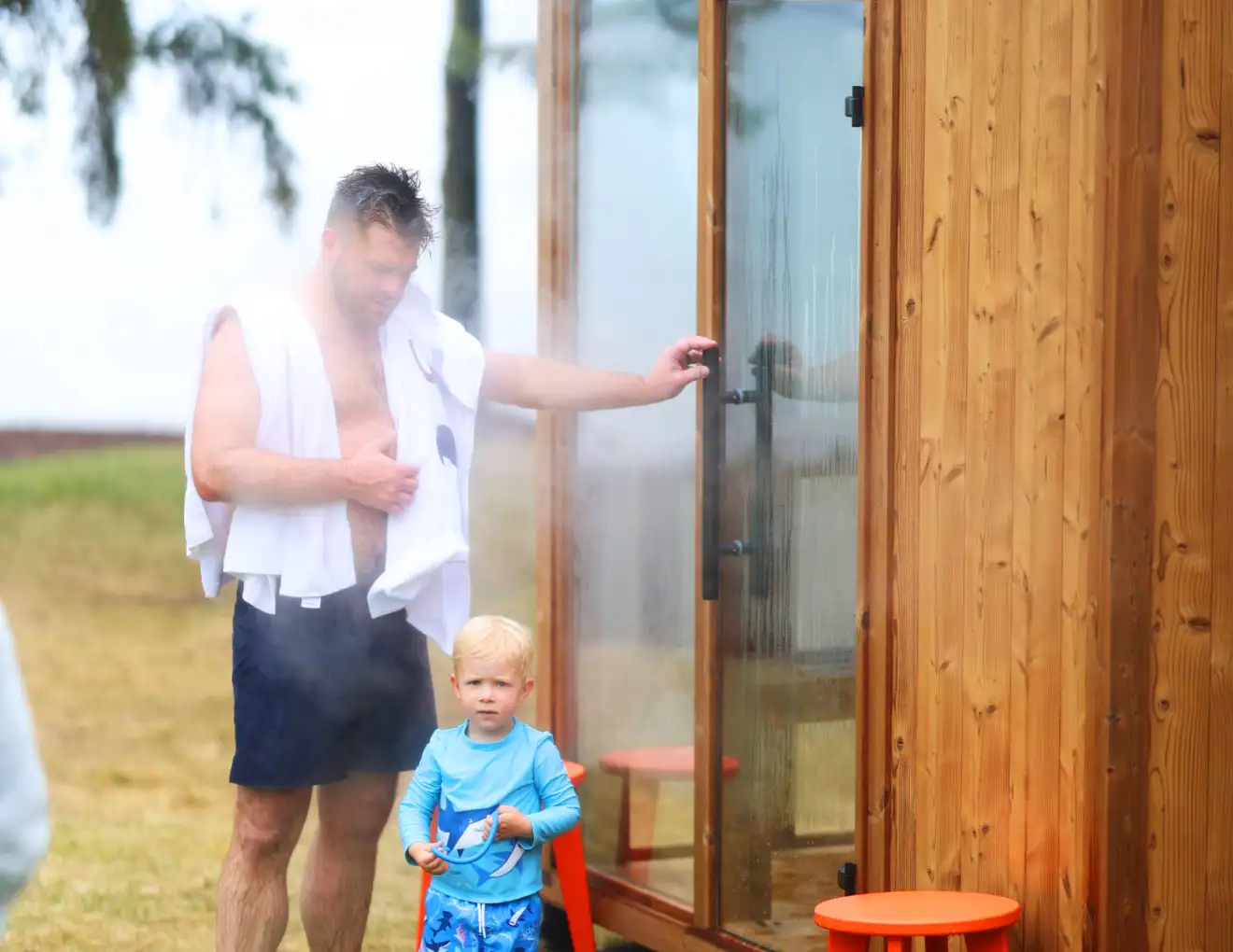 A man wears navy swim trunks with white towels over his shoulders, next to a small blonde boy in a light blue rash guard and trunks outside of a Traditional Mod Pod Sauna.